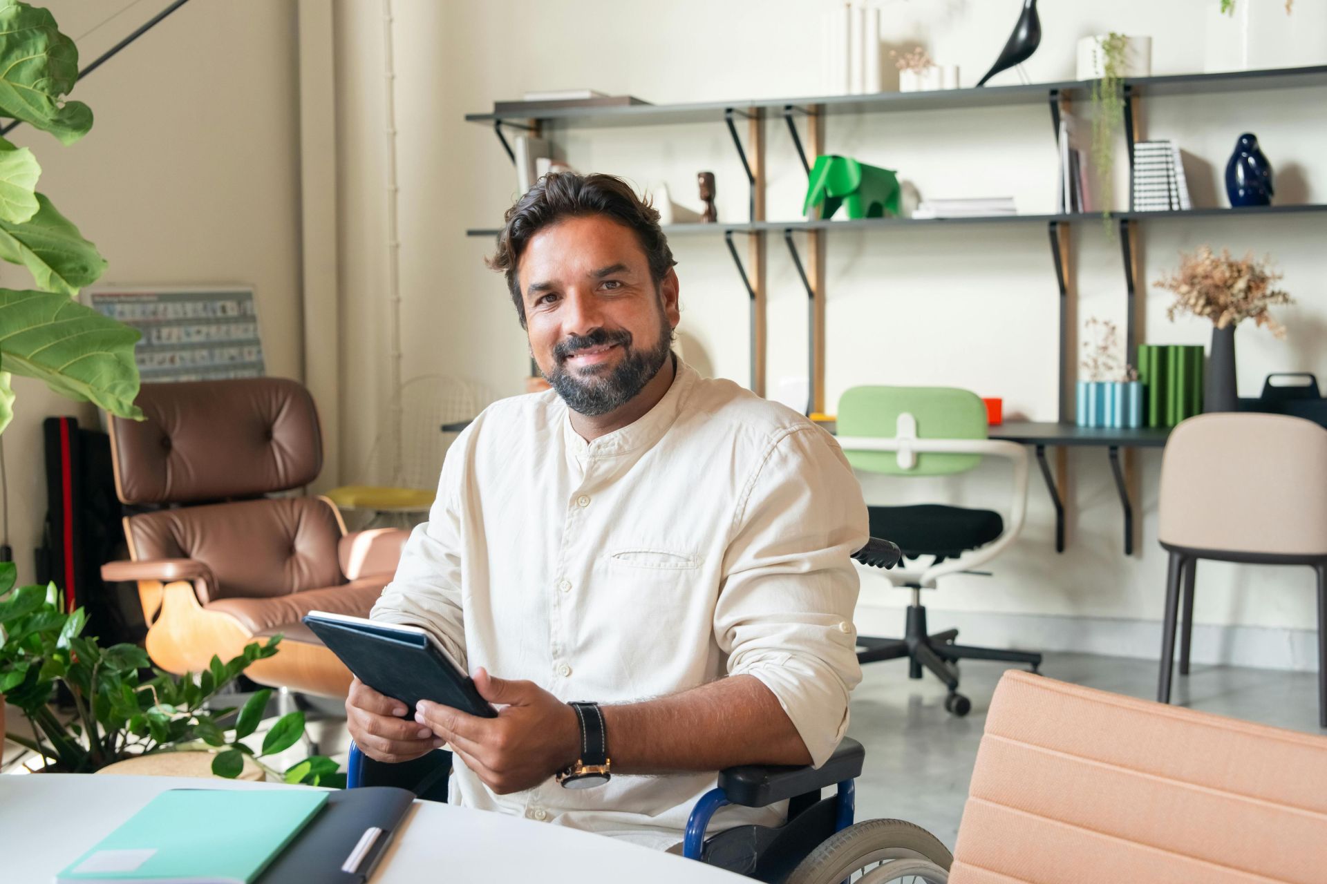 Bearded man in a wheelchair using a tablet and smiling indoors in a modern office.